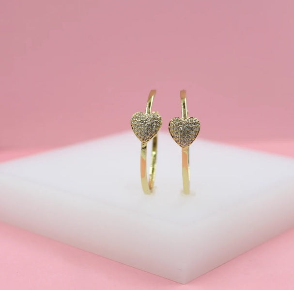 A pair of gold hoop earrings featuring pave-set crystal heart charms rests on a white display stand against a pink background.