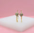 A pair of gold hoop earrings featuring pave-set crystal heart charms rests on a white display stand against a pink background.
