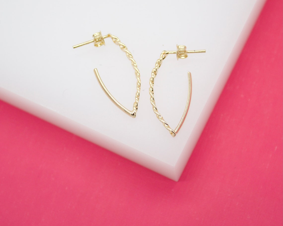 A pair of minimalist gold hoop earrings featuring a unique twisted design rests on a white and pink display surface.