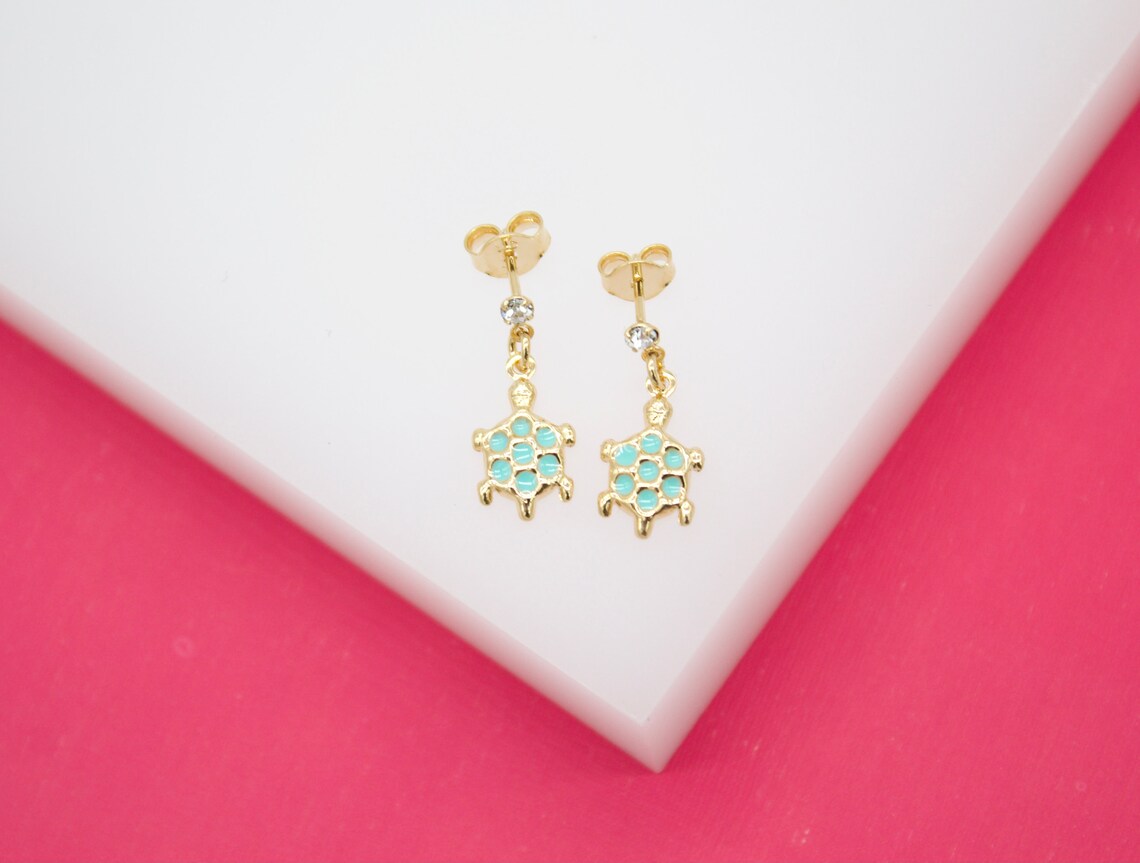 A pair of gold turtle stud earrings featuring turquoise enamel shells rests on a white surface against a bright pink background.