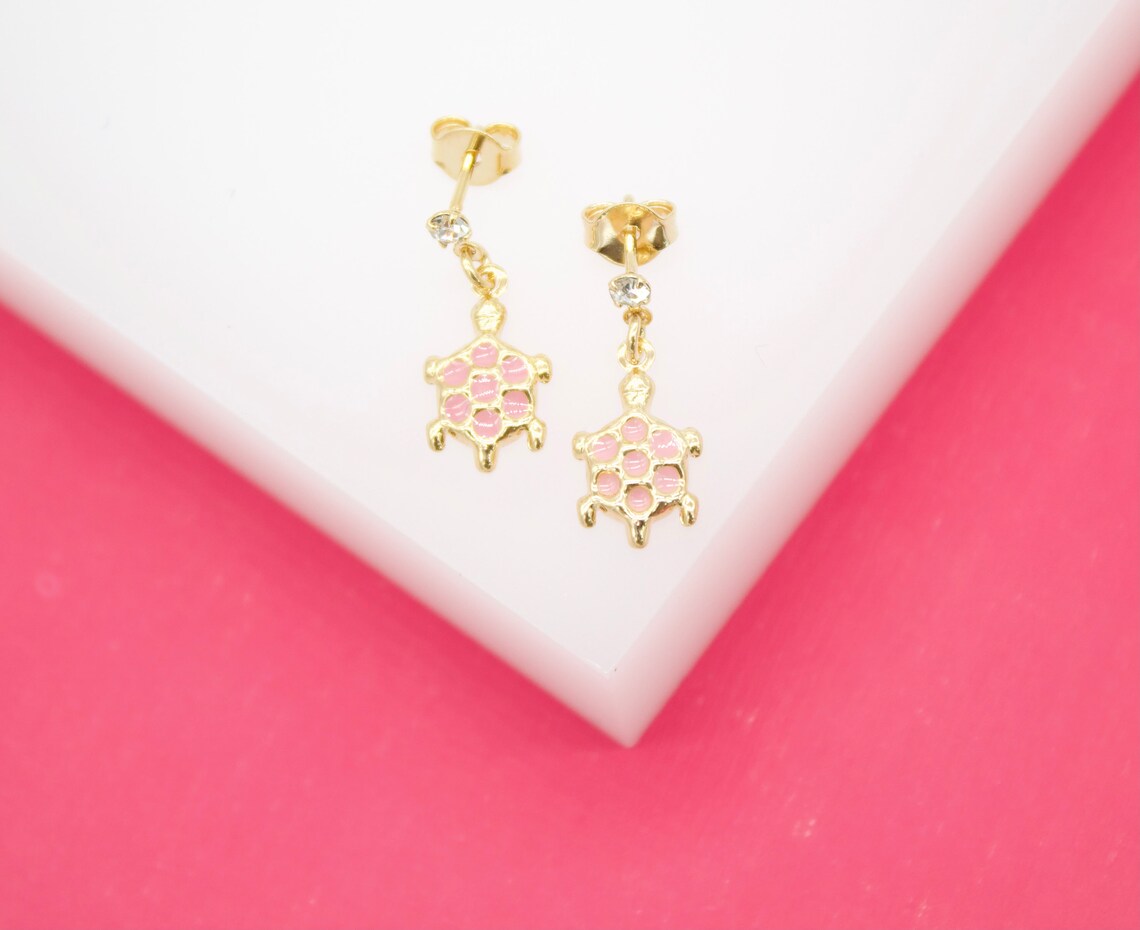 A pair of gold turtle stud earrings featuring pink enamel shells and sparkling crystal accents rest on a white surface against a bright pink background.