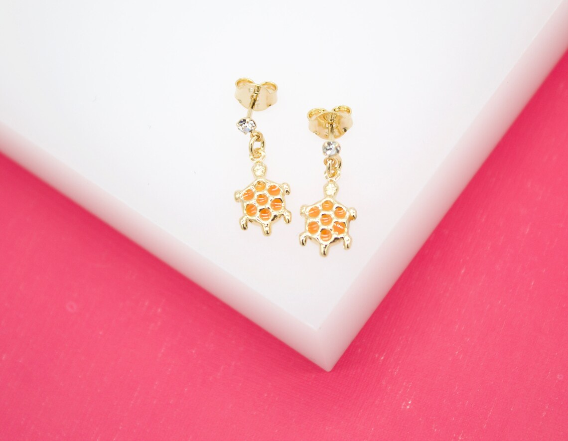 A pair of gold turtle earrings with orange shells and sparkling crystal accents rest on a white display surface against a vibrant pink background.
