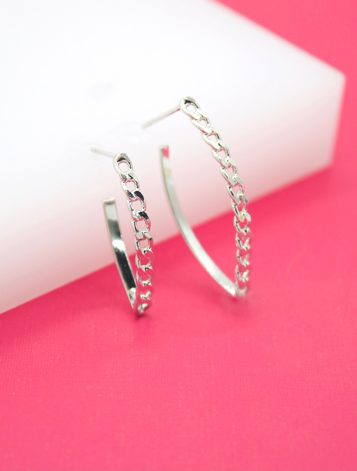 A pair of silver chain link hoop earrings rests on a pink and white background.
