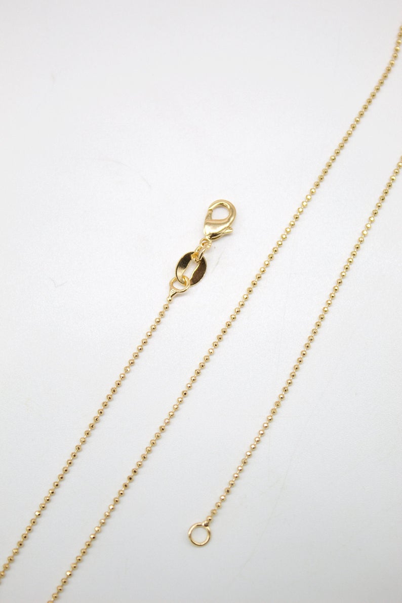 A delicate gold ball chain necklace rests on a white surface, highlighting its clasp and fine beaded links.