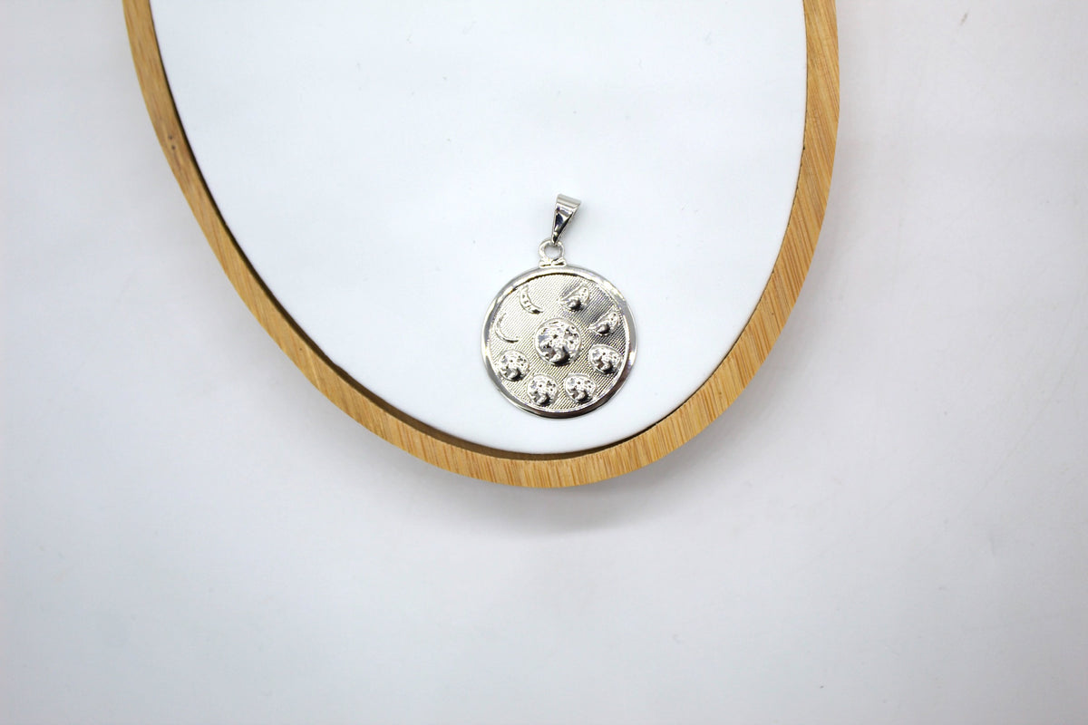 A silver medallion pendant featuring a celestial pattern rests on a white display board with a wooden frame.
