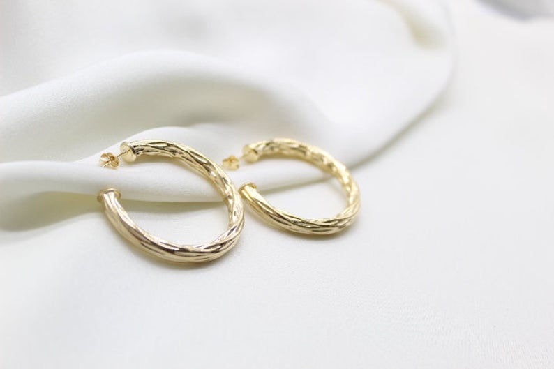A pair of textured gold hoop earrings rests elegantly on soft white silk fabric.