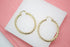 A pair of polished gold twist hoop earrings rests on a white display against a pink background.