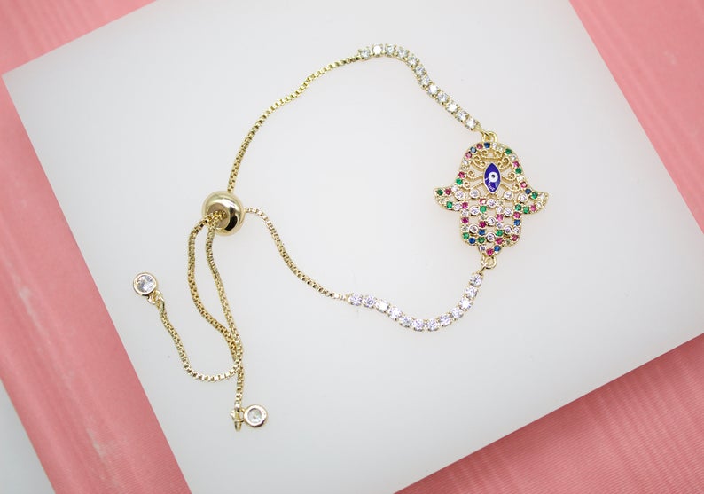 A delicate gold Hamsa hand bracelet features sparkling multicolored stones and a central blue evil eye charm against a neutral background.