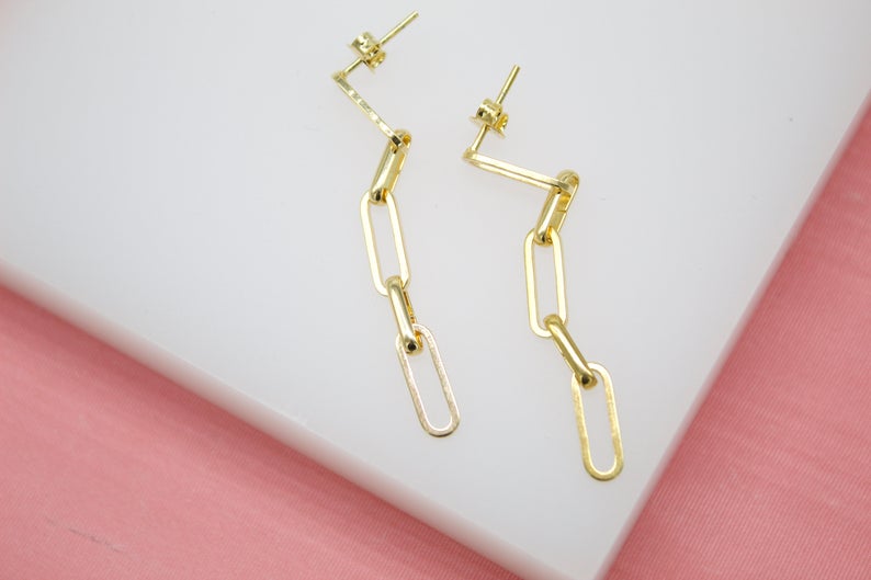 A pair of gold paperclip chain drop earrings rests on a white and pink surface.