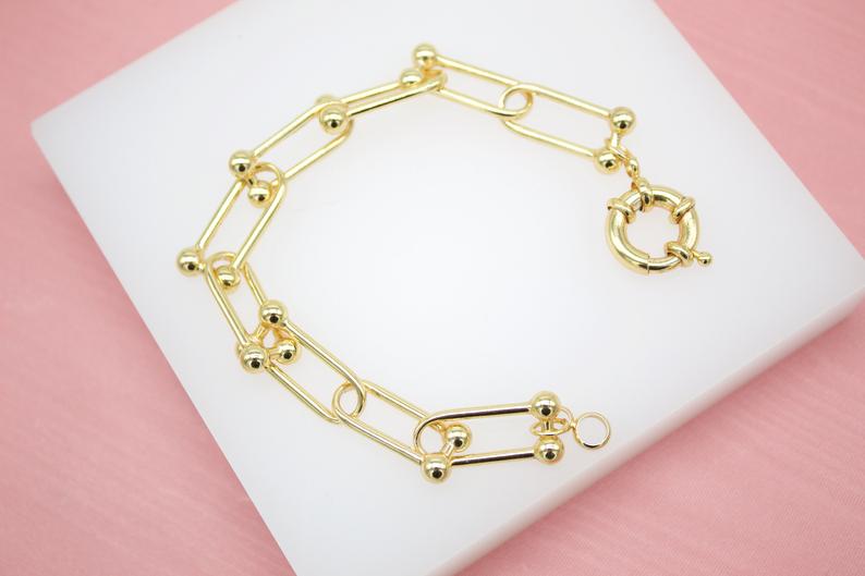 A polished gold paperclip link bracelet with round ball accents rests on a white display surface.