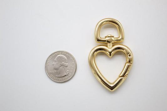A shiny gold heart-shaped swivel lobster clasp rests next to a US quarter coin for scale on a white background.