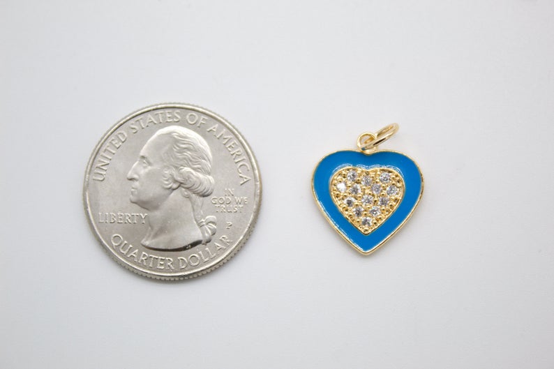 A blue and gold heart-shaped charm with a pavé diamond center rests beside a US quarter for size comparison.