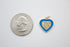 A blue and gold heart-shaped charm with a pavé diamond center rests beside a US quarter for size comparison.