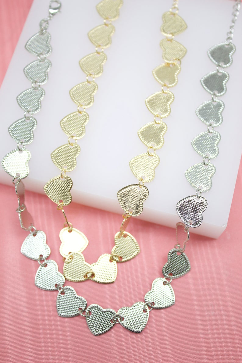Four metallic heart-link necklaces, in gold and silver, rest on a white display against a pink background.