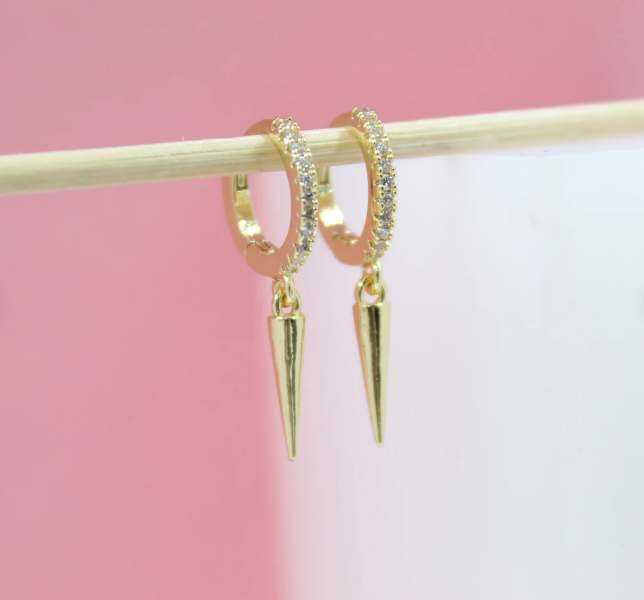 Two gold hoop earrings featuring sparkling pave-set stones and hanging spike charms hang from a wooden stick against a pink and white background.