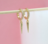 A pair of gold spike huggie earrings featuring sparkling pave crystals hangs from a wooden stick against a soft pink background.