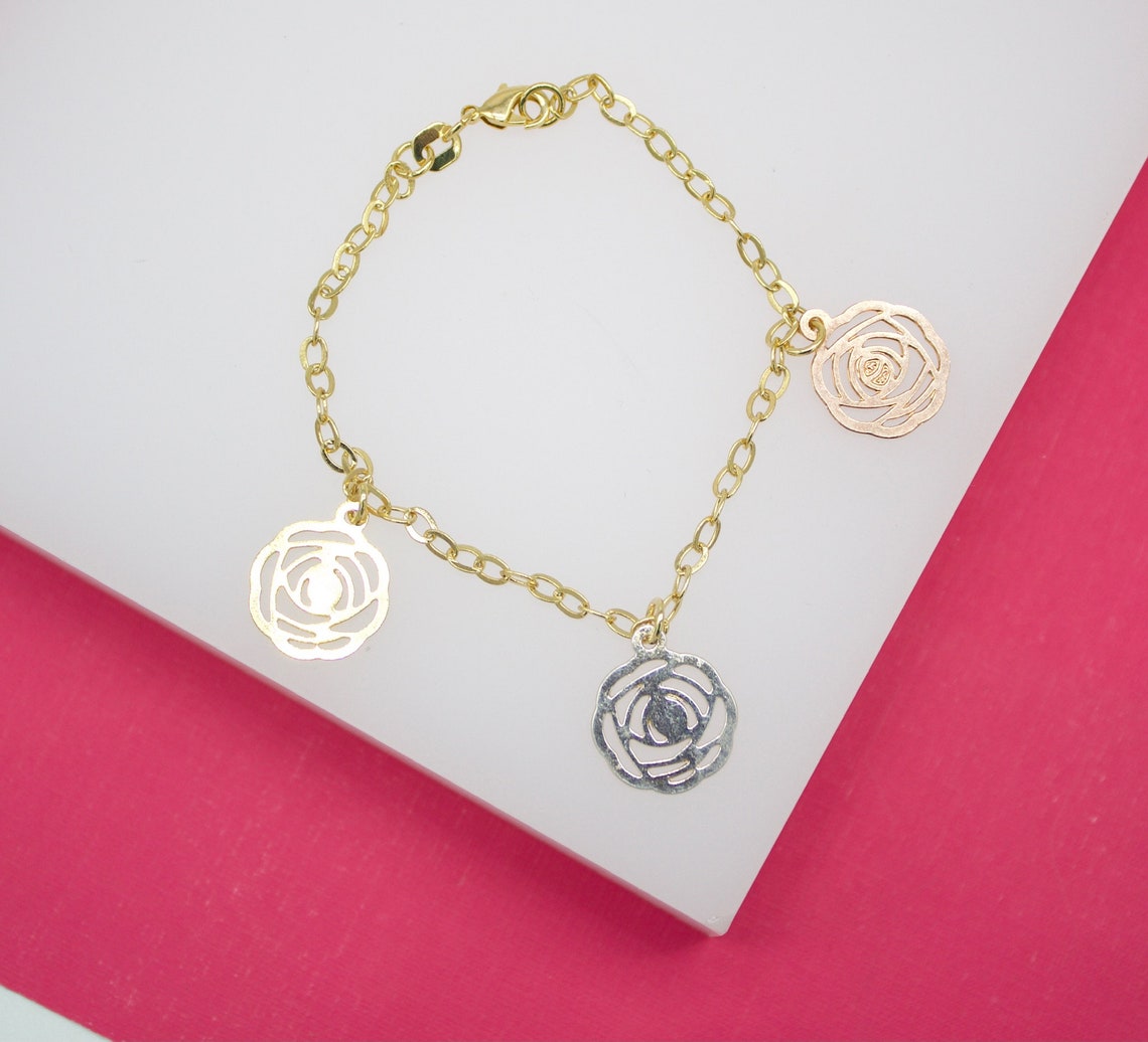 A delicate gold link charm bracelet displays three cutout rose flower charms in yellow, white, and rose gold against a white and pink background.