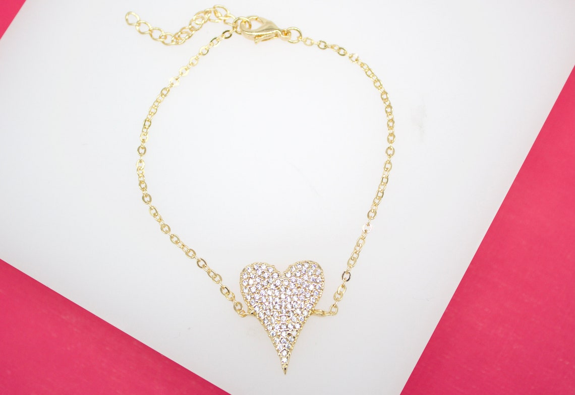 A gold chain bracelet features a sparkling, pave diamond-encrusted heart pendant centered on a white display surface.
