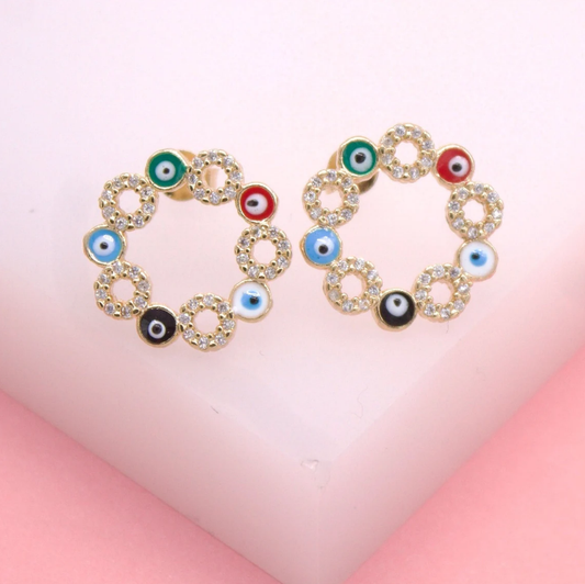 A pair of gold evil eye stud earrings, encrusted with cubic zirconia and featuring multicolored enamel charms, rests on a pink display surface.