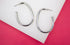 A pair of sleek silver hoop earrings rests on a white and pink background.