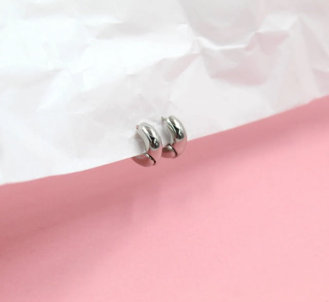 A pair of polished silver hoop earrings rest on a textured white paper backdrop against a soft pink surface.