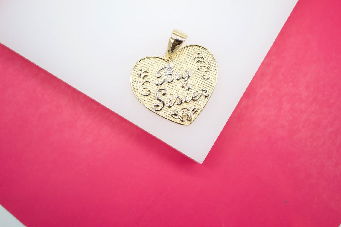 A gold heart-shaped pendant featuring the engraved words "Big Sister" rests on a white display surface against a vibrant pink background.