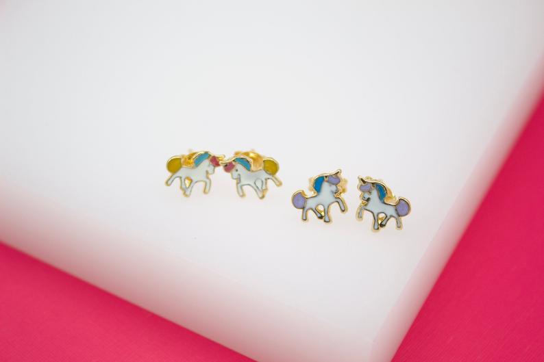 Two pairs of colorful unicorn earrings rest on a white display surface against a pink background.