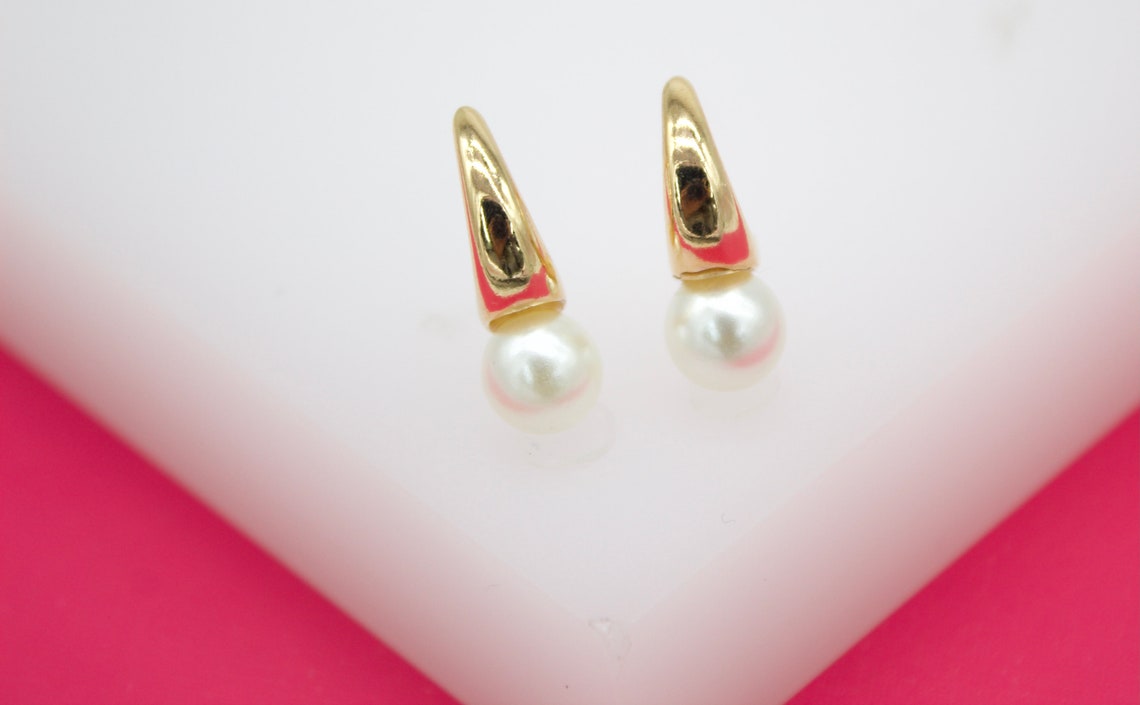 A pair of gold earrings featuring dangling white pearls rests on a white and pink display stand.