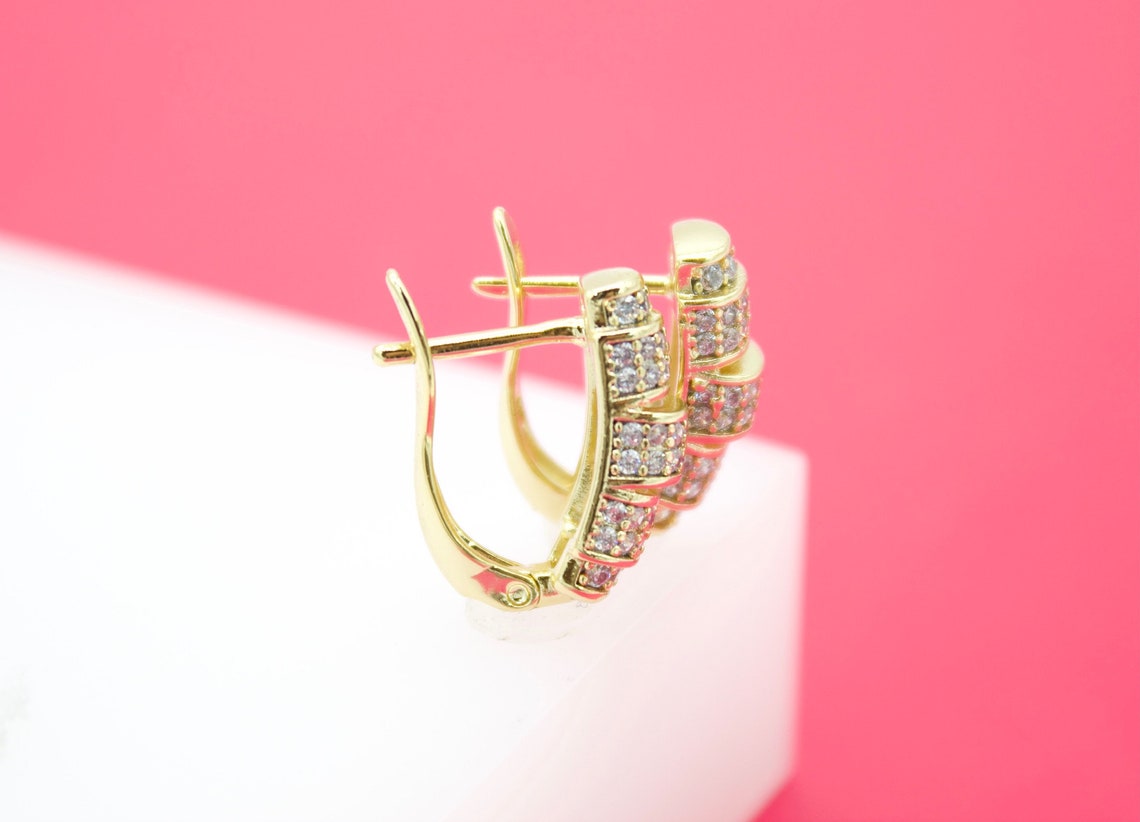 A pair of gold hoop earrings embellished with sparkling CZ stones rests on a white display stand against a pink background.