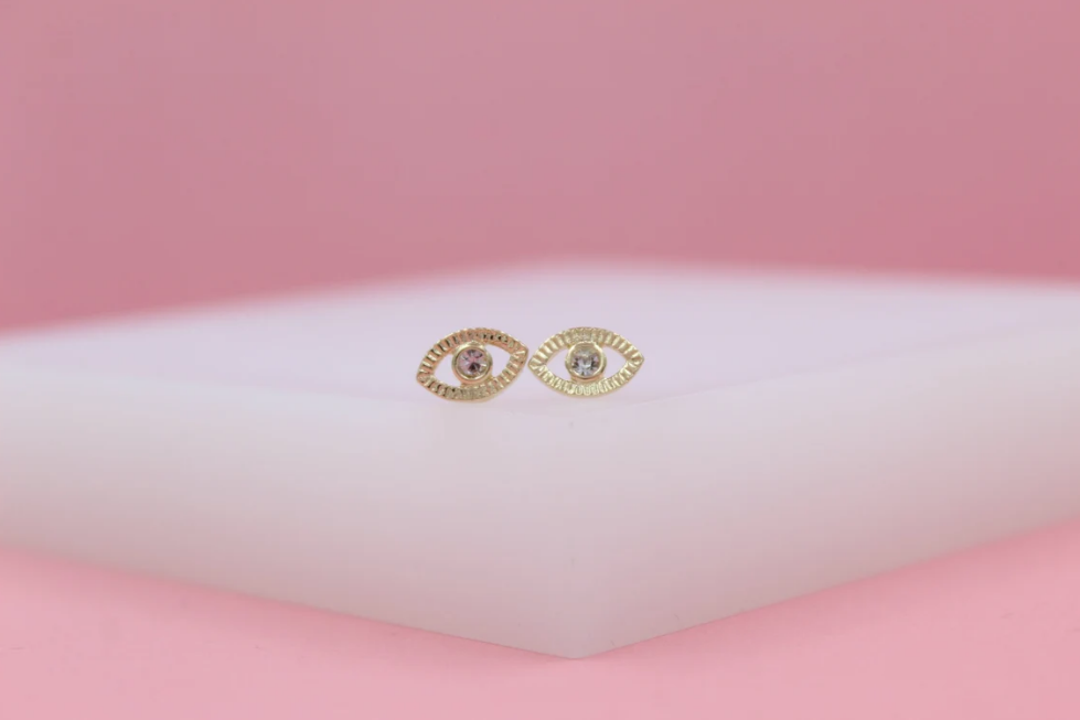 Two gold evil eye stud earrings with crystal centers rest on a white display against a pink background.