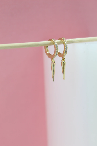 A pair of gold huggie hoop earrings with sparkling cubic zirconia pavé and dangling spike charms hangs from a wooden stick against a soft pink and white background.