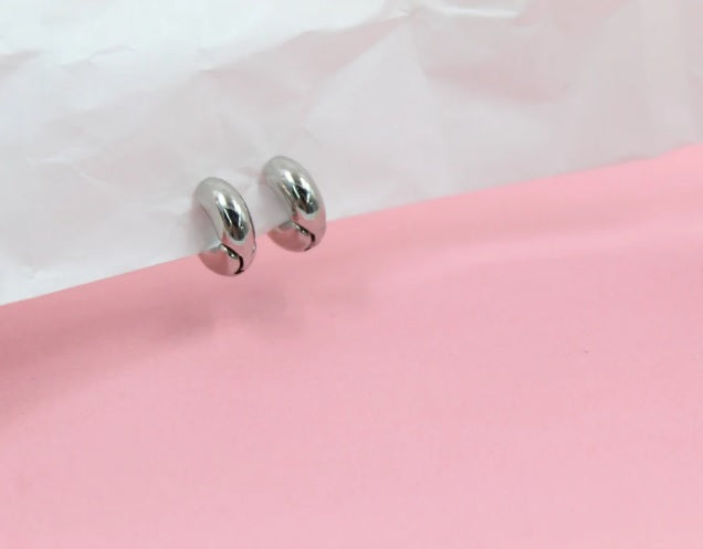 Two shiny silver hoop earrings rest on a white crinkled paper against a soft pink background.