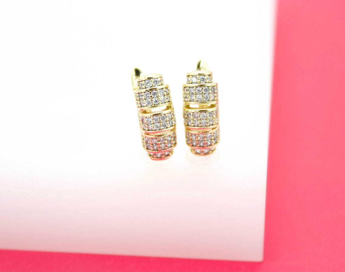 A pair of gold huggie hoop earrings encrusted with sparkling cubic zirconia stones rests on a white display against a pink background.