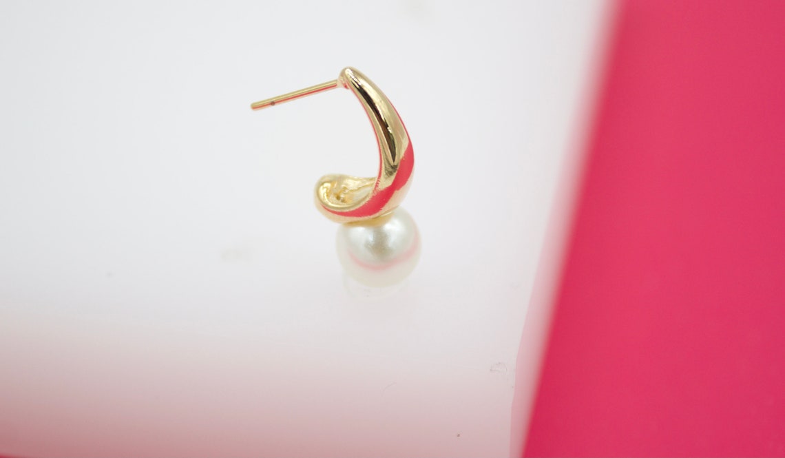 A gold and pink enamel hoop earring accented with a dangling white pearl rests against a white and pink background.