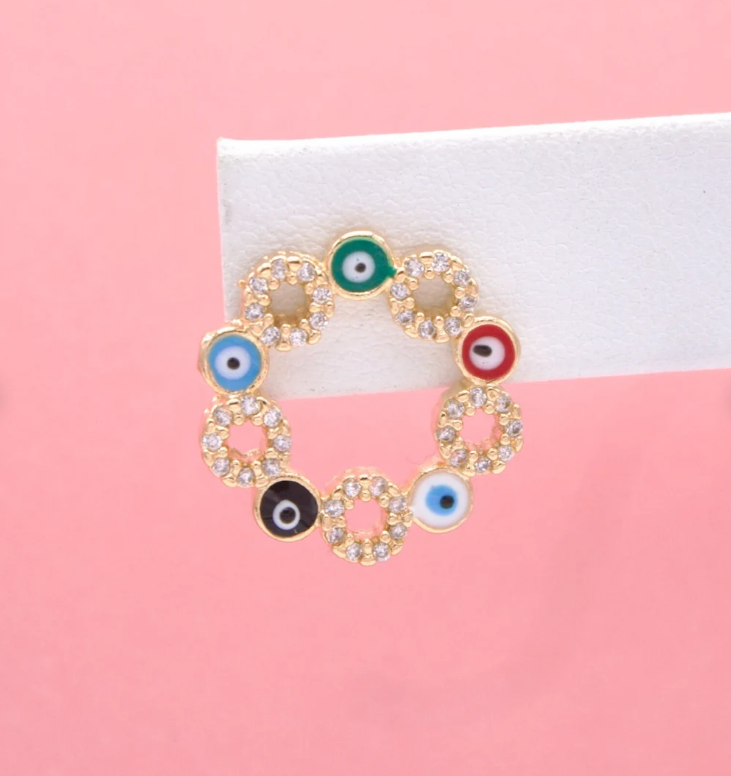 A gold evil eye hoop earring adorned with multicolored enamel charms and sparkling cubic zirconia stones is displayed on a white earring card against a soft pink background.