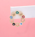 A gold evil eye hoop earring adorned with multicolored enamel charms and sparkling cubic zirconia stones is displayed on a white earring card against a soft pink background.