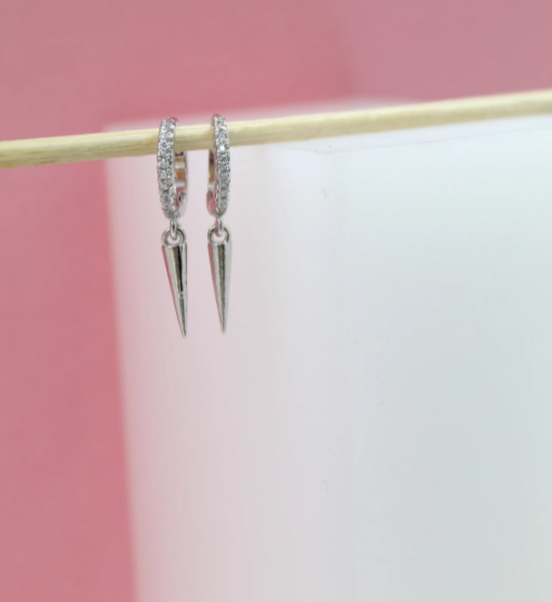 A pair of dangling pavé hoop spike earrings hangs from a wooden stick against a white and pink backdrop.