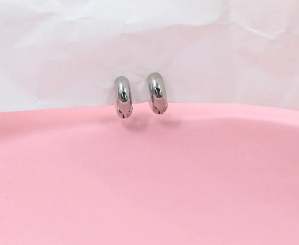 Two sleek, silver hoop earrings rest elegantly on a soft pink surface against a white paper background.