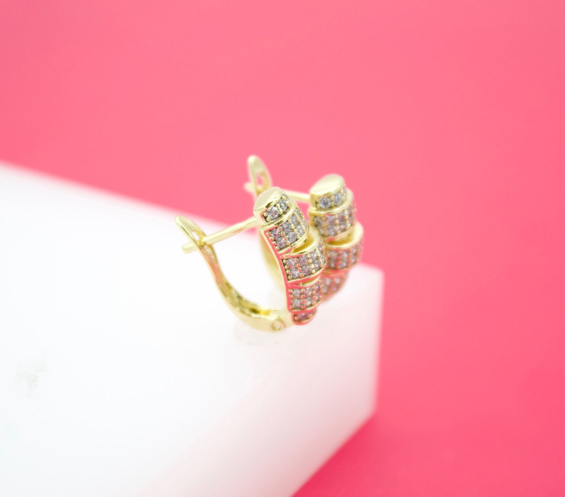 A pair of ornate gold hoop earrings encrusted with sparkling cubic zirconia stones rests on a white display block against a vibrant pink background.