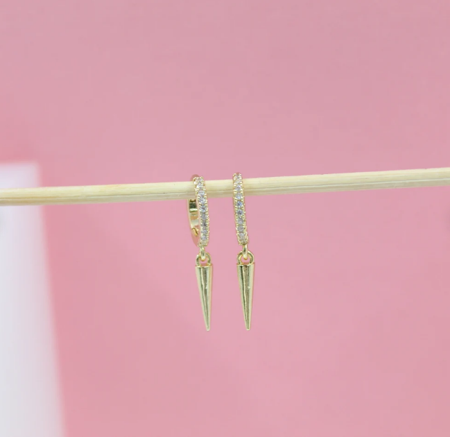 Two gold hoop earrings featuring crystal-embellished bands and dangling spike charms hang from a wooden stick against a soft pink background.