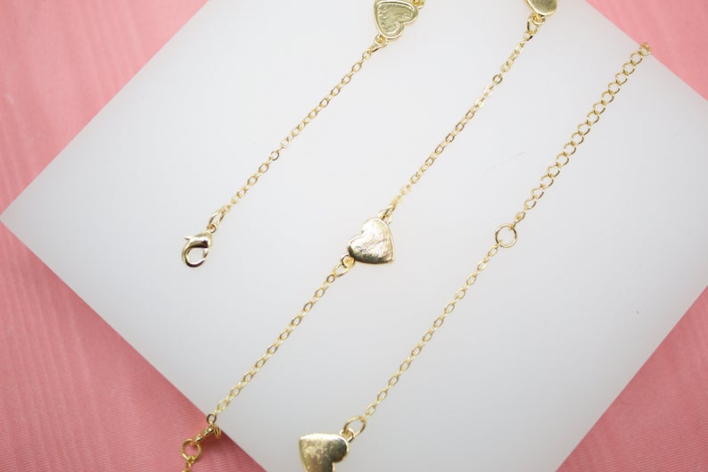 A delicate gold heart-shaped charm anklet rests on a white display surface against a pink background.