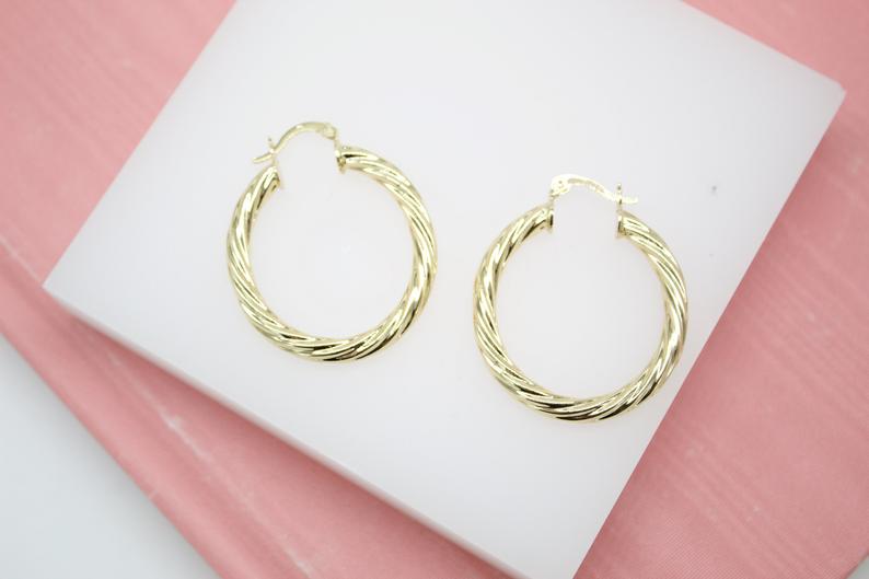 A pair of polished gold twist hoop earrings rests on a white display surface against a pink background.
