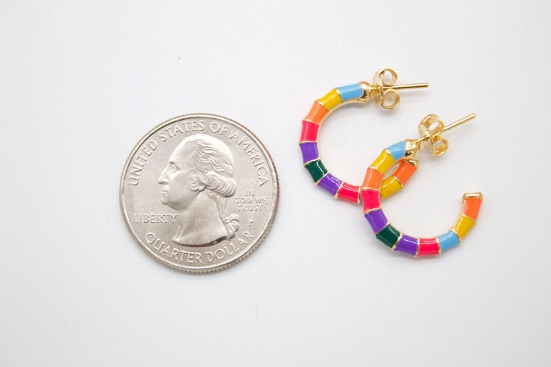 A pair of colorful enameled gold hoop earrings lies next to a US quarter coin for scale on a white surface.