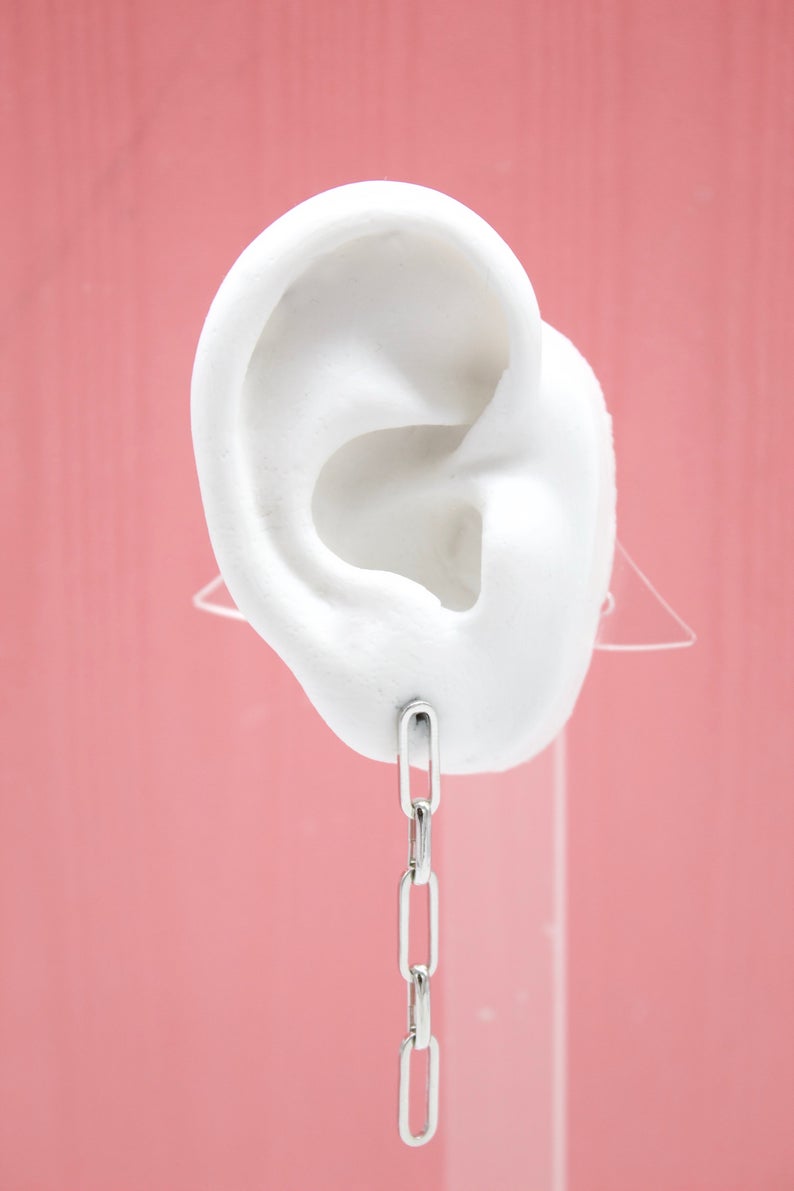 A white display ear showcases a dangling silver chain link earring against a pink background.