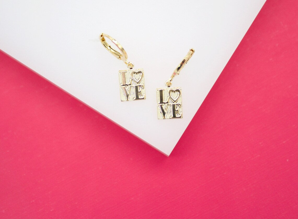 A pair of gold huggie hoop earrings featuring rectangular charms with the word "LOVE" engraved on them rests against a white and pink backdrop.