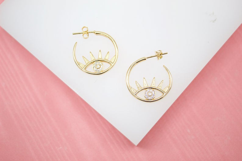 A pair of gold hoop earrings featuring an evil eye design with sparkling crystal accents rests on a white display against a pink background.