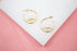 A pair of gold hoop earrings featuring an evil eye design with sparkling crystal accents rests on a white display against a pink background.