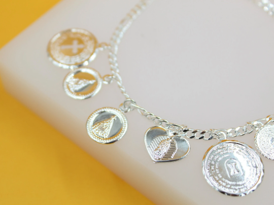 A delicate silver charm bracelet displays religious medallions and a heart pendant on a white and yellow display surface.