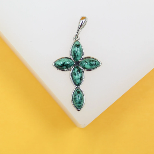 A silver cross pendant featuring marbled green turquoise stones rests on a white and yellow display stand.