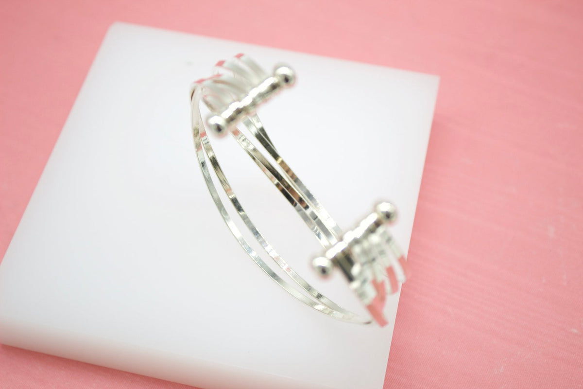 A shiny silver multi-band cuff bracelet rests on a white square display tile against a soft pink background.